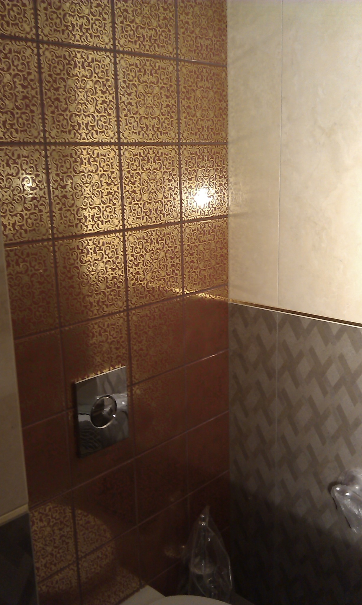 Wykończona ściana łazienki z brązowo-złotymi płytkami o wzorzystej fakturze, sąsiadująca z jasnymi, gładkimi płytkami i fragmentem ściany z szarymi płytkami w geometryczny wzór. Widoczny przycisk...