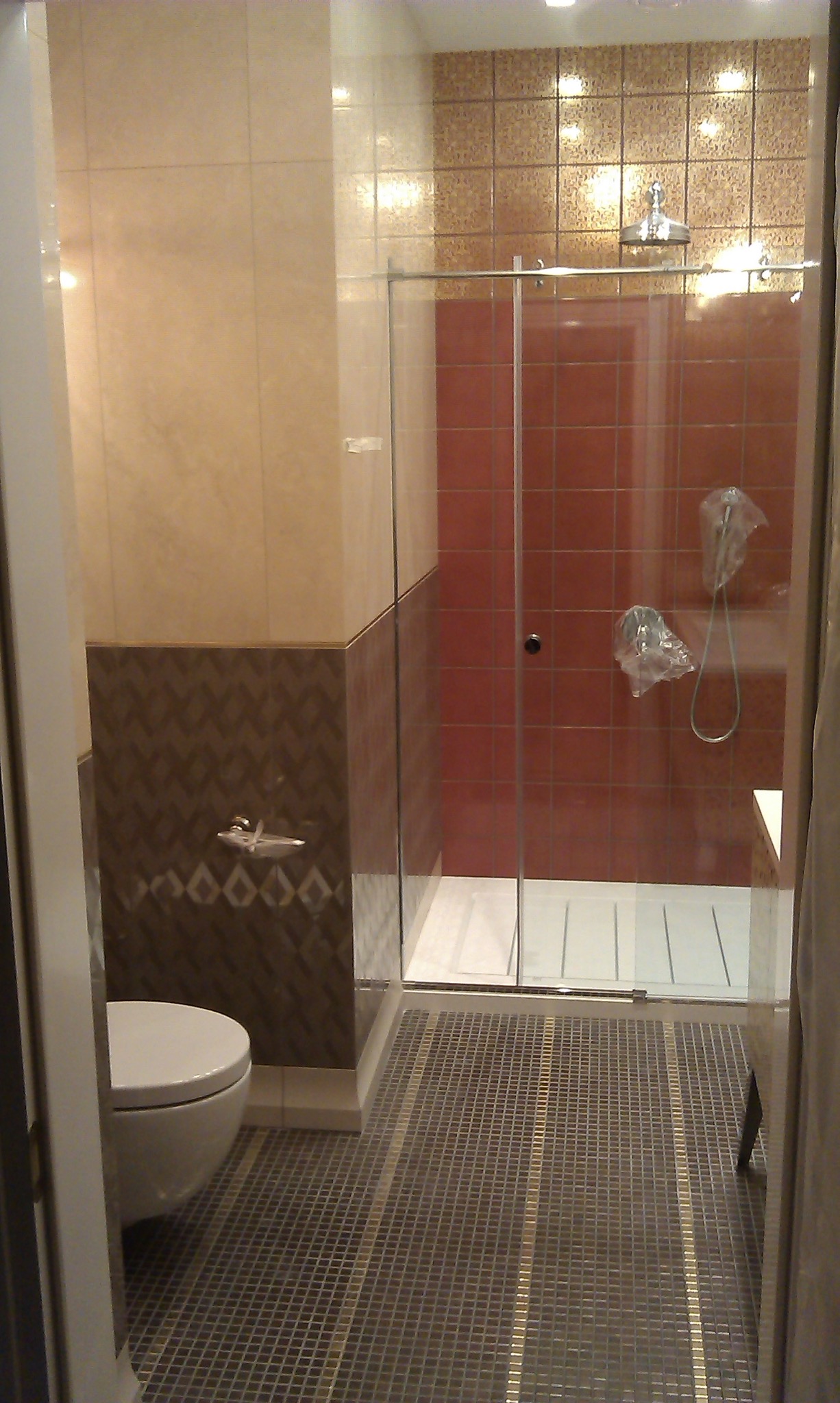 Wykończona łazienka z kabiną prysznicową typu walk-in, brązowo-beżowe płytki na ścianach, mozaika na podłodze z złotymi paskami, toaleta z boku, słuchawka prysznicowa w folii ochronnej.