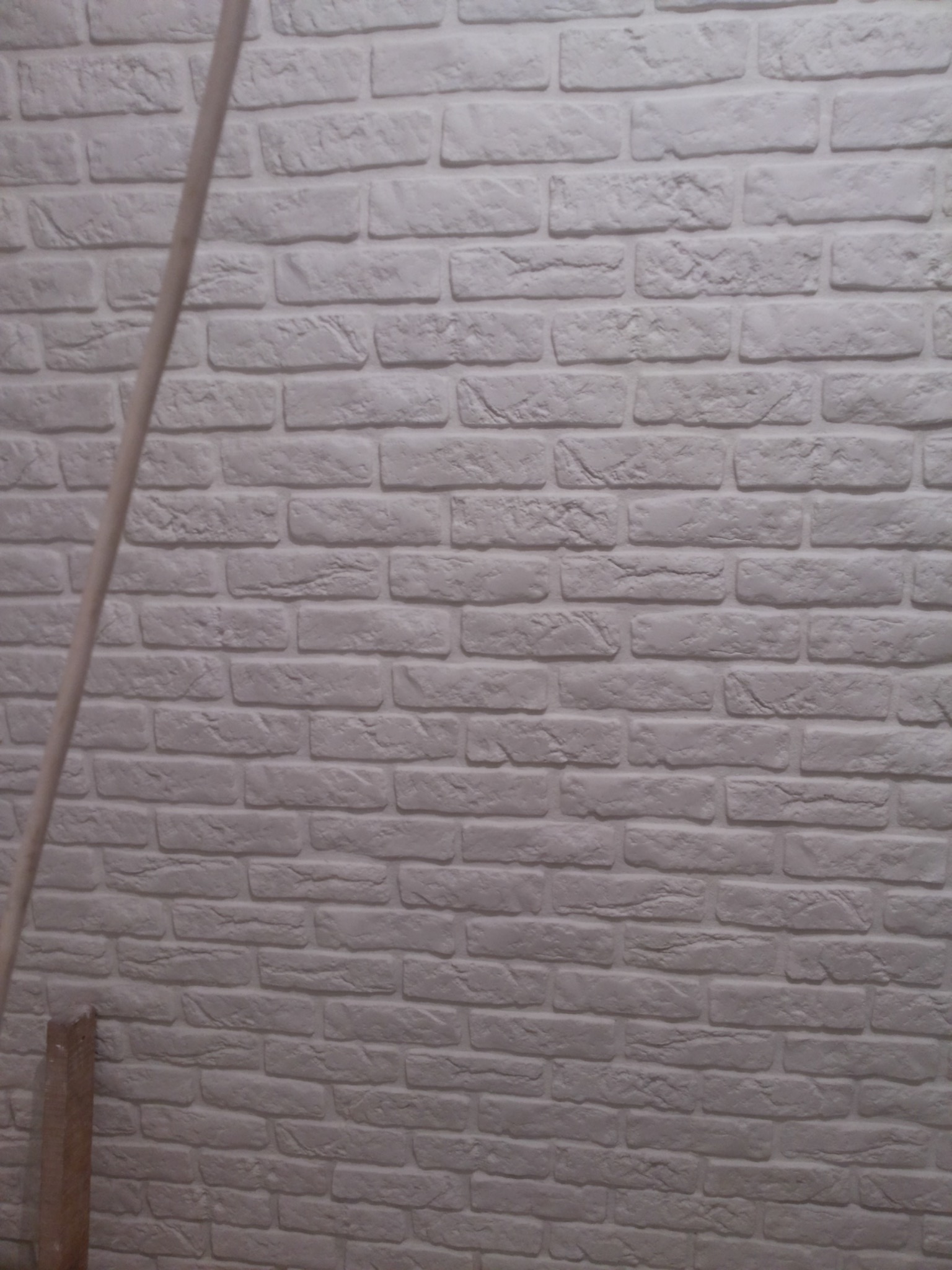 Biała ściana z cegły dekoracyjnej, widoczny fragment drewnianej podpory w lewym dolnym rogu, nierówne fugi.