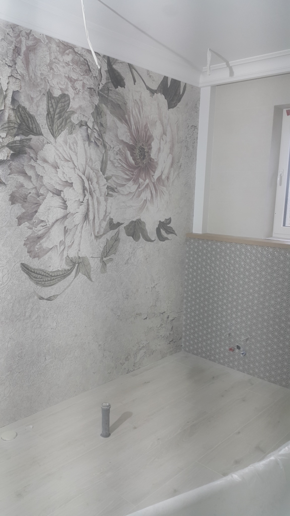 Ściana łazienki z fototapetą w kwiaty i fragmentem ściany wyłożonej szarą mozaiką geometryczną; podłoga wyłożona jasnymi panelami imitującymi drewno, widoczna rura odpływowa.