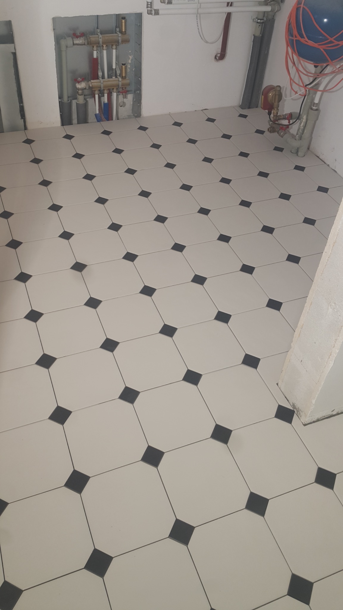 Ułożona podłoga z jasnych, kwadratowych płytek z ciemnymi, kwadratowymi wstawkami w narożach, widoczne instalacje grzewcze na ścianie.