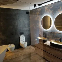 Nowoczesna łazienka z czarno-złotymi umywalkami nablatowymi, podświetlanymi okrągłymi lustrami, ciemnymi płytkami imitującymi kamień i drewnopodobną podłogą, z widoczną wanną wolnostojącą...