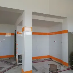 Wnętrze w trakcie układania płytek: białe płytki ścienne z poziomym pasem pomarańczowych płytek, widoczne kable na podłodze, krzesło i grzejnik.