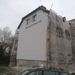 PPHU Mag-Bud - Budynek mieszkalny w trakcie prac elewacyjnych, częściowo zakryty siatką ochronną z nadrukiem reklamowym farb Caparol, widoczne rusztowania i napis MAGAYZNOWA 108 na świeżo pomalowanej ścianie.