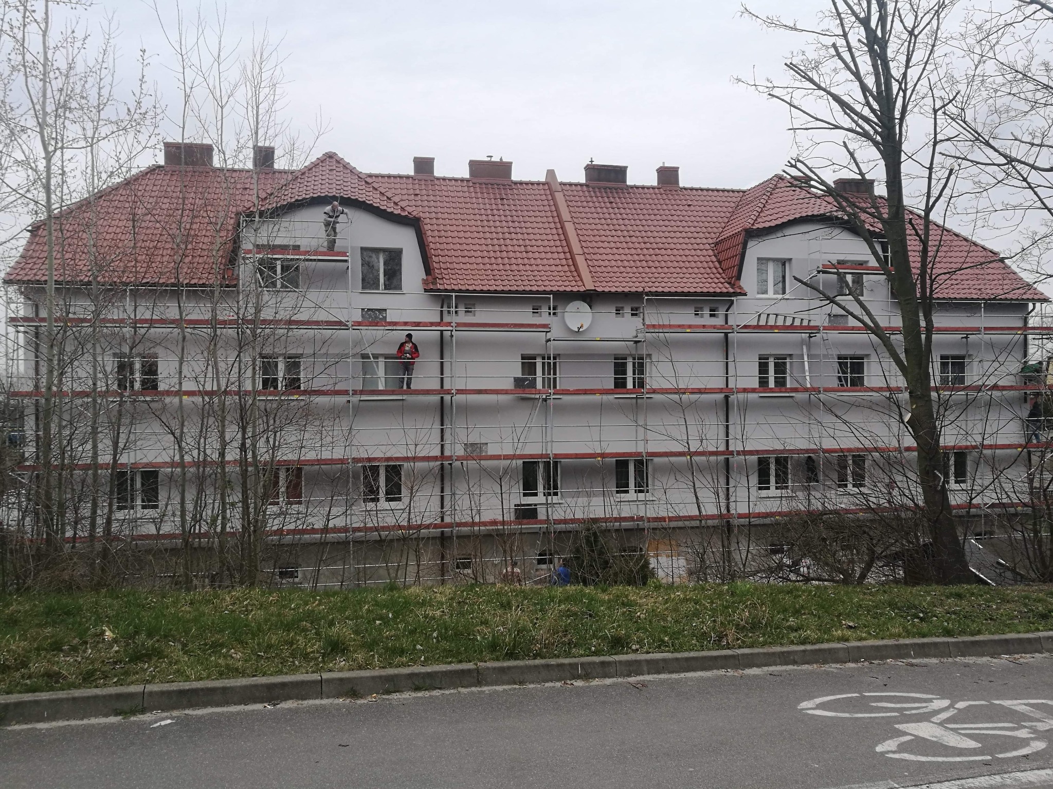 Wielorodzinny budynek w trakcie malowania elewacji, widoczne rusztowania i pracownik w czerwonej kurtce na drugim piętrze, czerwona dachówka, pochmurne niebo.
