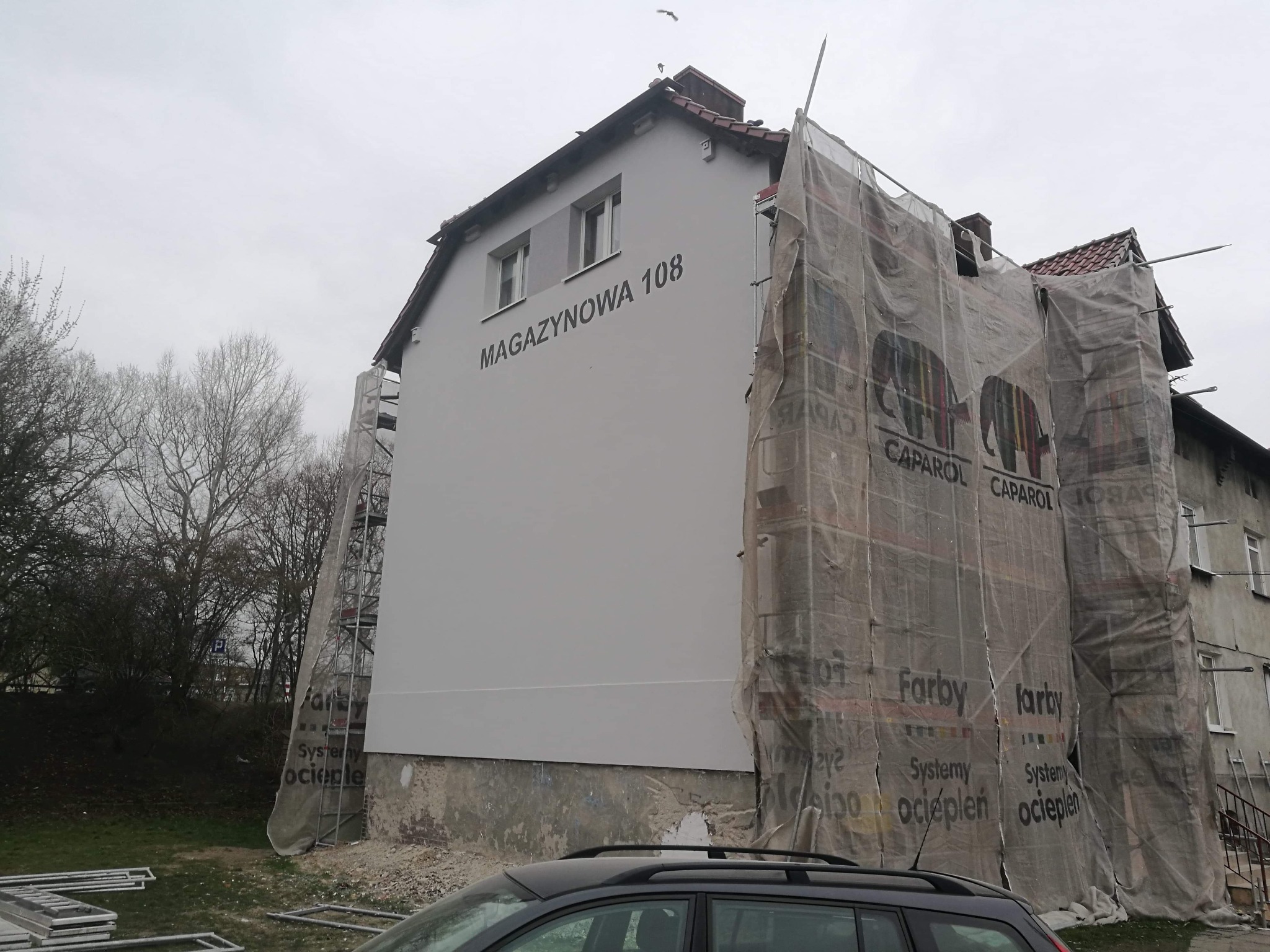 Budynek mieszkalny w trakcie prac elewacyjnych, częściowo zakryty siatką ochronną z nadrukiem reklamowym farb Caparol, widoczne rusztowania i napis MAGAYZNOWA 108 na świeżo pomalowanej ścianie.