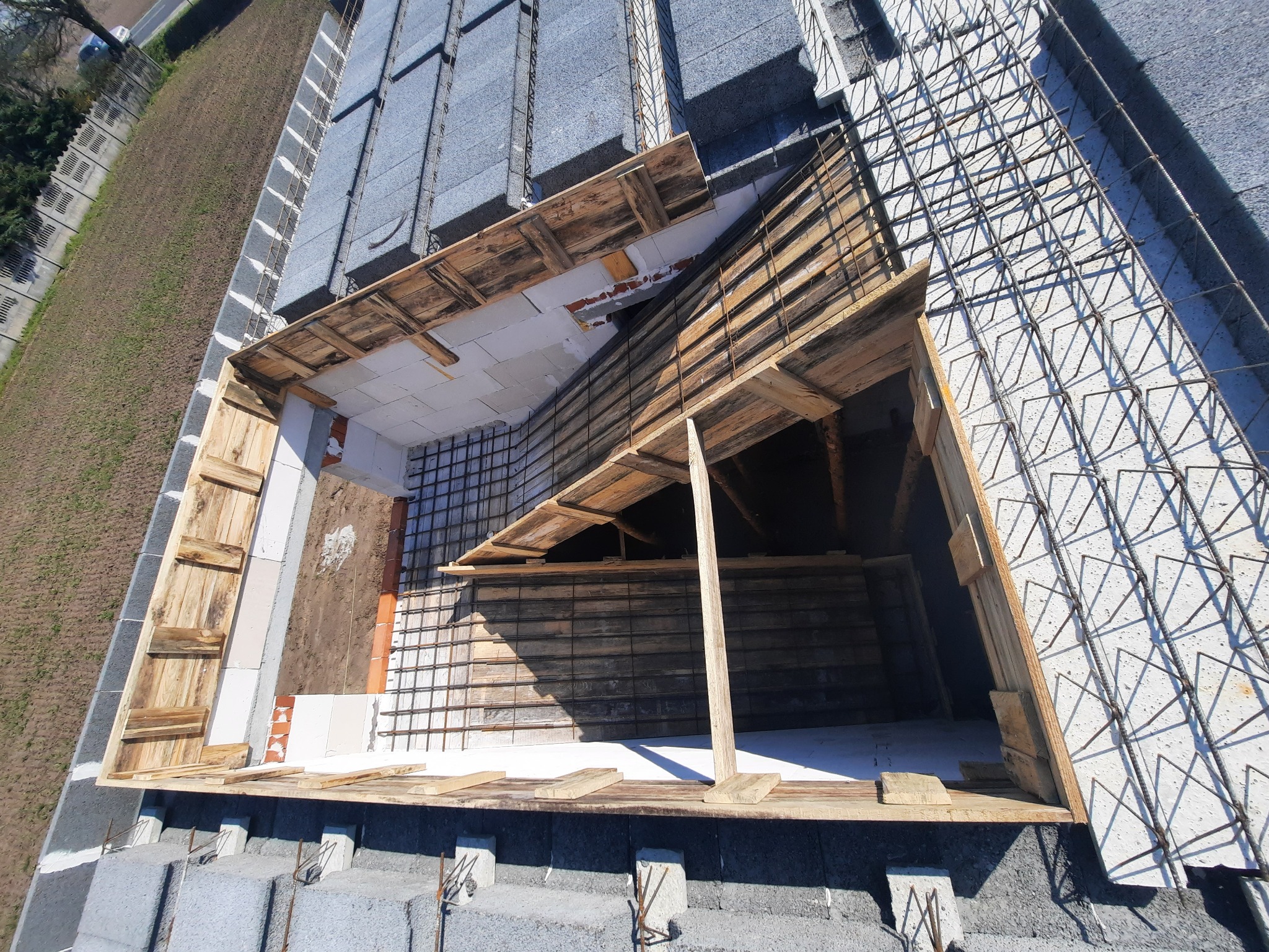Konstrukcja poddasza w trakcie budowy z widocznym szkieletem drewnianym, siatką zbrojeniową i bloczkami betonowymi, ujęcie z góry w słoneczny dzień.