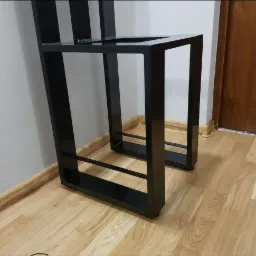 Część metalowej konstrukcji krzesła barowego z czarnych profili kwadratowych, widoczna na tle drewnianej podłogi i białej ściany.