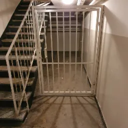 Biała, metalowa krata zabezpieczająca dostęp do przestrzeni pod schodami w budynku mieszkalnym, widoczne rury pod sufitem i schody z białą balustradą.