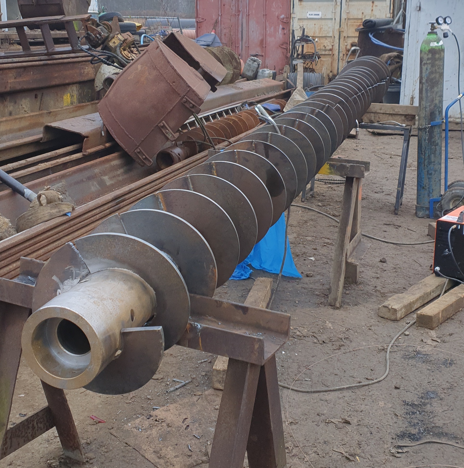 Śruba Archimedesa z przyspawanymi dyskami, wsparta na metalowych kozłach w warsztacie, widoczny spawacz i butla z gazem.