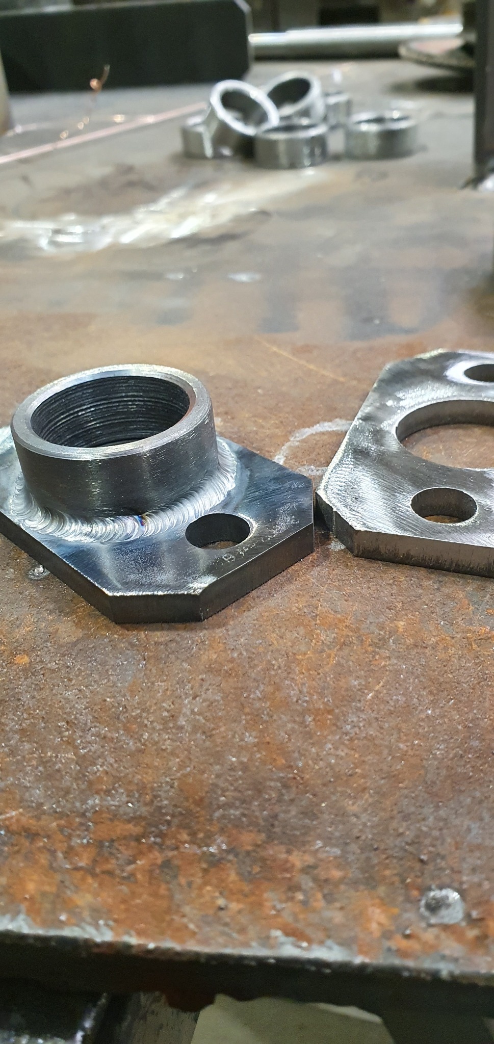 Wyszlifowane, spawane elementy stalowe leżące na zardzewiałym stole warsztatowym, widoczne ślady spawania i obróbki metalu.