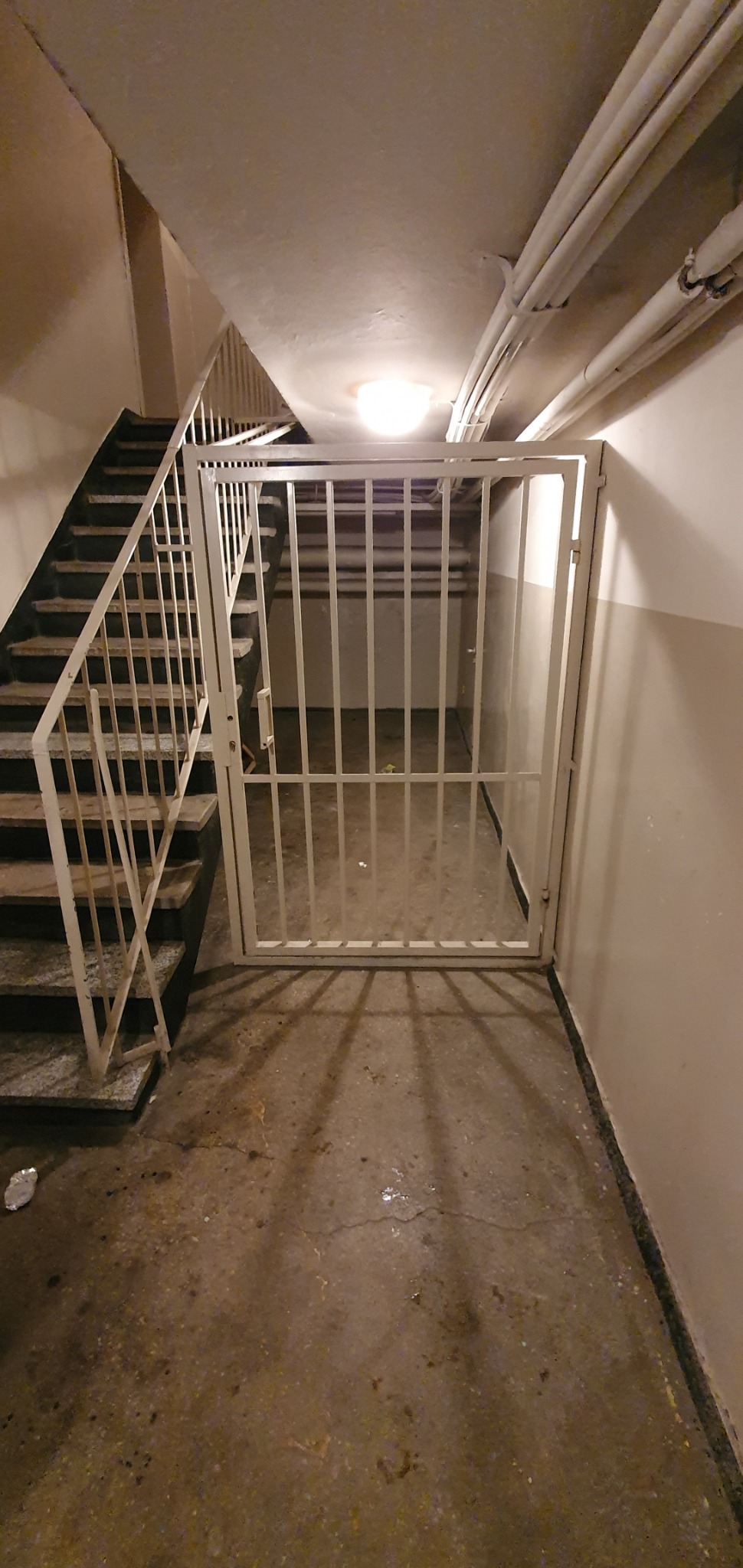 Biała, metalowa krata zabezpieczająca dostęp do przestrzeni pod schodami w budynku mieszkalnym, widoczne rury pod sufitem i schody z białą balustradą.