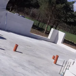 Wylany fundament z wystającymi rurami kanalizacyjnymi, ściany z białych bloczków w tle, widok na las i pole.