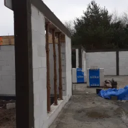Wznoszenie ścian z betonu komórkowego na fundamencie z izolacją, widoczne drewniane stemple podtrzymujące nadproże, w tle palety z bloczkami Silka.
