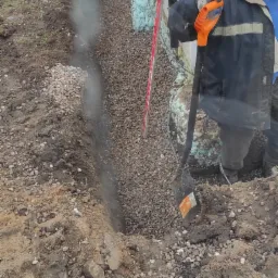 Praca ziemne przy fundamencie budynku: wykop wypełniony żwirem, zabezpieczona ściana fundamentowa z widoczną izolacją termiczną, pracownik z łopatą o pomarańczowym trzonku.