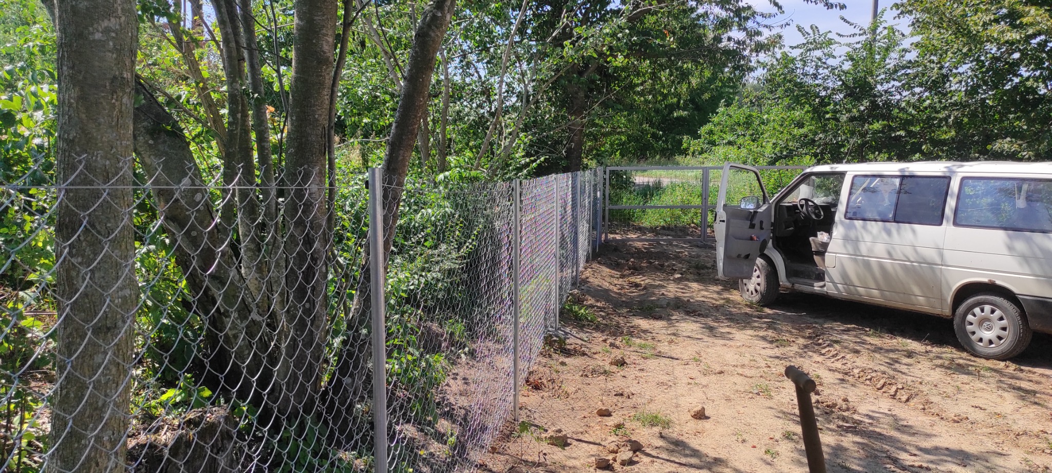 Montaż ogrodzenia z siatki ocynkowanej pomiędzy drzewami, z otwartą furtką i zaparkowanym białym busem z otwartymi drzwiami.