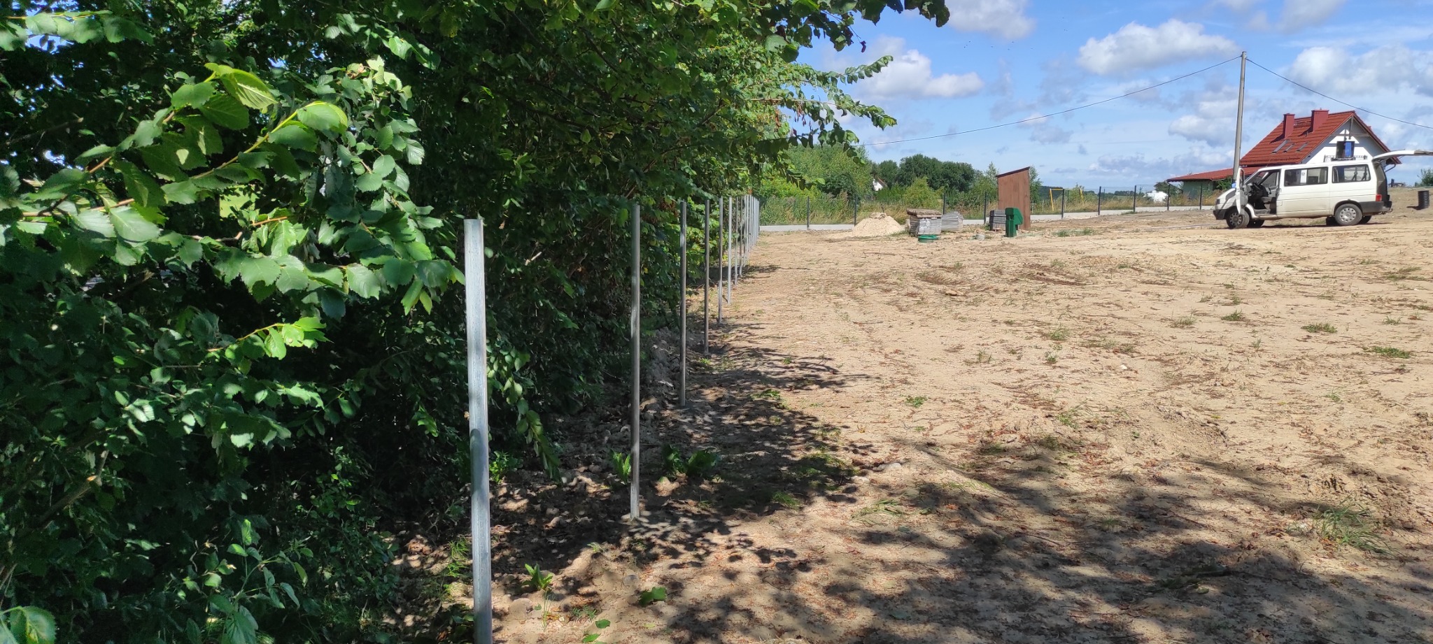 Montaż słupków ogrodzeniowych wzdłuż linii drzew na piaszczystym terenie, widoczny samochód dostawczy i dom w tle.