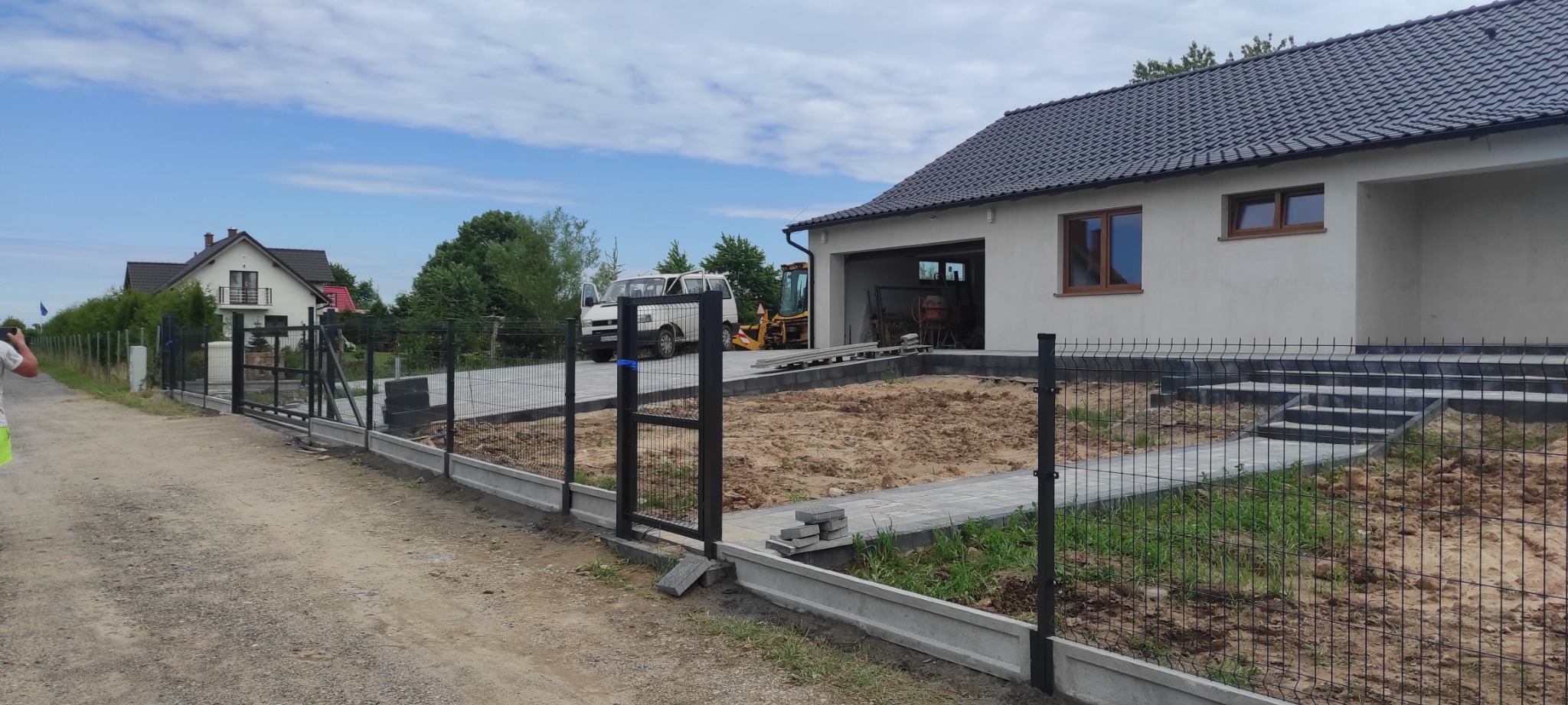 Montaż ogrodzenia panelowego z podmurówką betonową na posesji z nowym domem, widoczny częściowo ułożony chodnik z kostki, koparka i bus w tle.