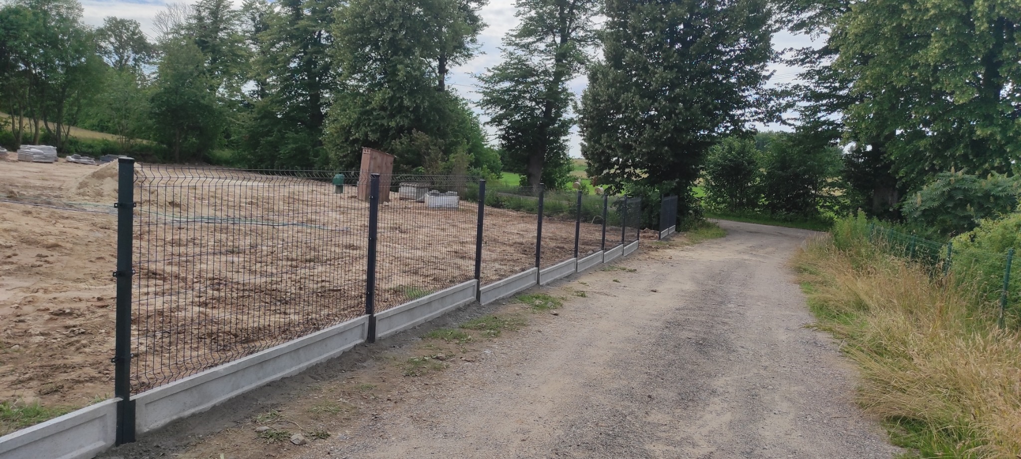 Nowoczesne ogrodzenie panelowe z podmurówką betonową na działce z przygotowanym terenem pod budowę, widok z drogi gruntowej.