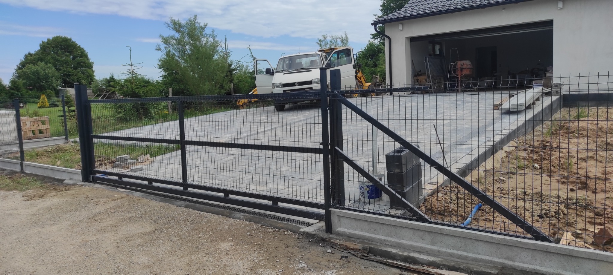 Czarna, przesuwna brama panelowa z siatki na betonowej podmurówce, widoczny fragment posesji z podjazdem z kostki, samochód dostawczy z otwartymi drzwiami i mini koparka w tle.
