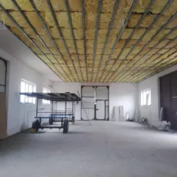 Wnętrze hali z widocznym ociepleniem sufitu wełną mineralną i metalową konstrukcją nośną, w tle wózek transportowy i materiały budowlane.