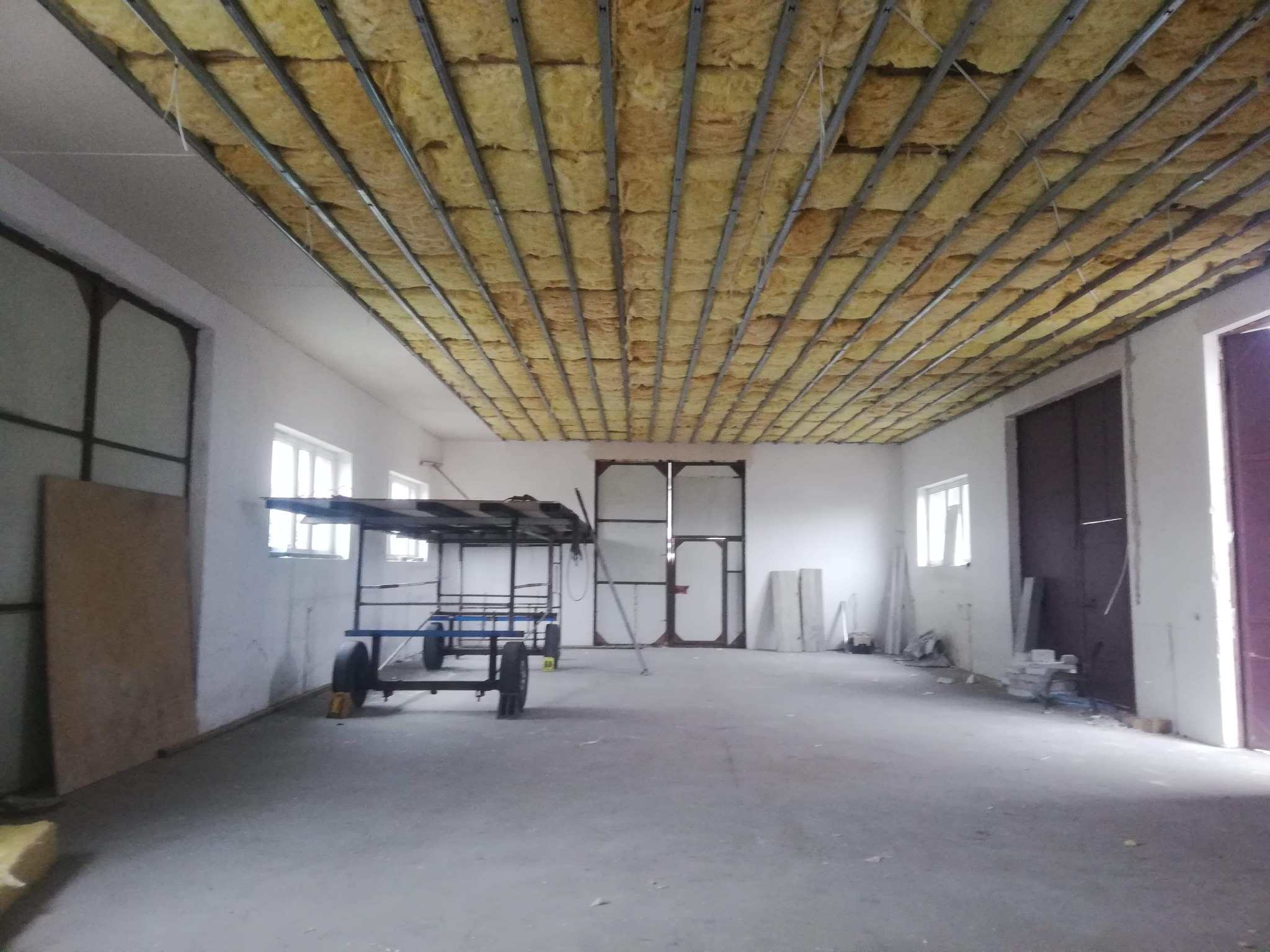 Wnętrze hali z widocznym ociepleniem sufitu wełną mineralną i metalową konstrukcją nośną, w tle wózek transportowy i materiały budowlane.