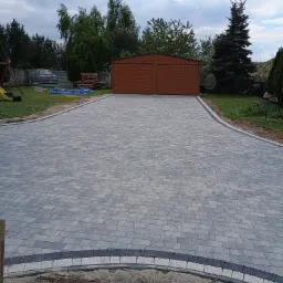 Szeroki podjazd z szarej kostki ułożonej we wzór jodełki, obrzeża z ciemnej kostki, w tle drewniany garaż i zieleń ogrodu, pochmurne niebo.