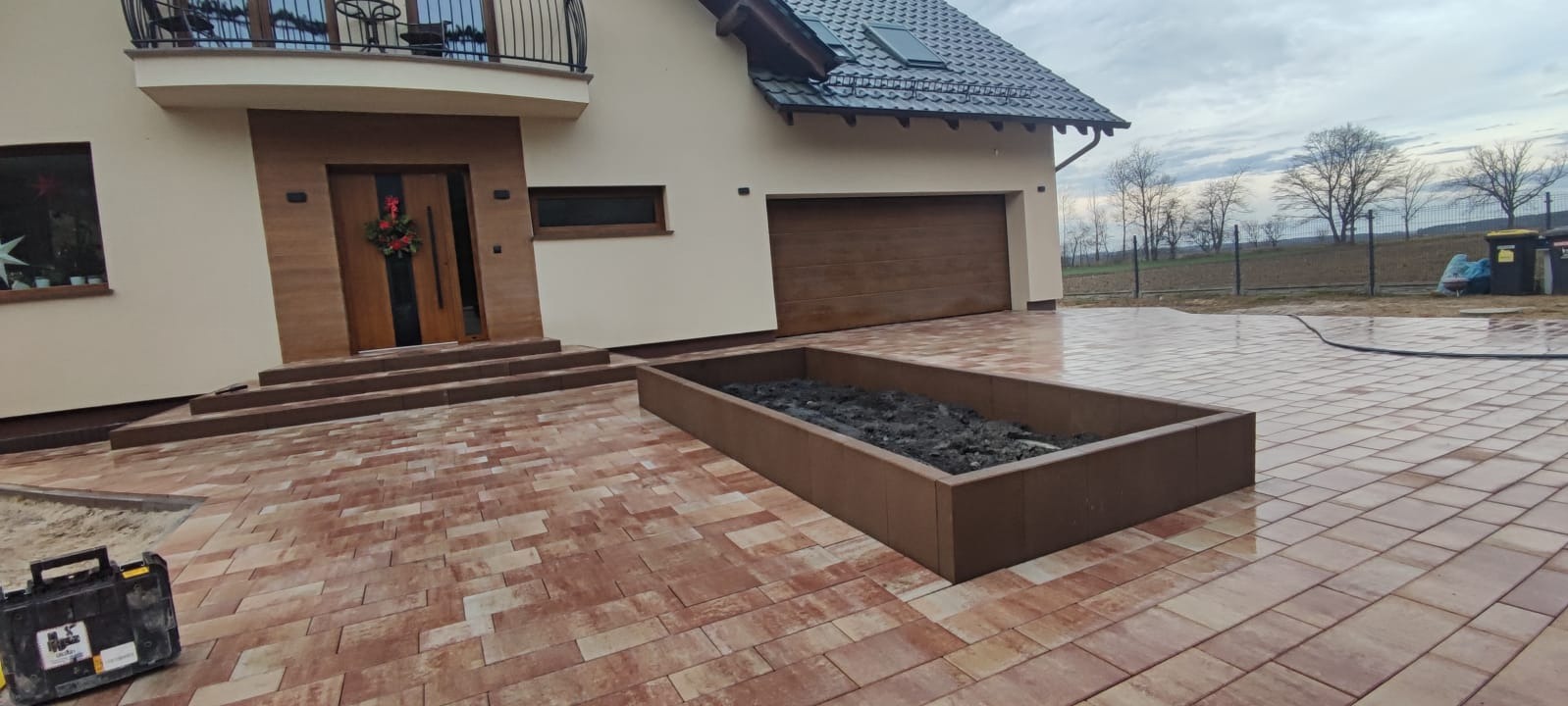Nowo ułożona, wzorzysta kostka brukowa w odcieniach czerwieni i beżu przed domem z brązowymi elementami, widoczna skrzynia na rośliny i skrzynka z narzędziami brukarskim.