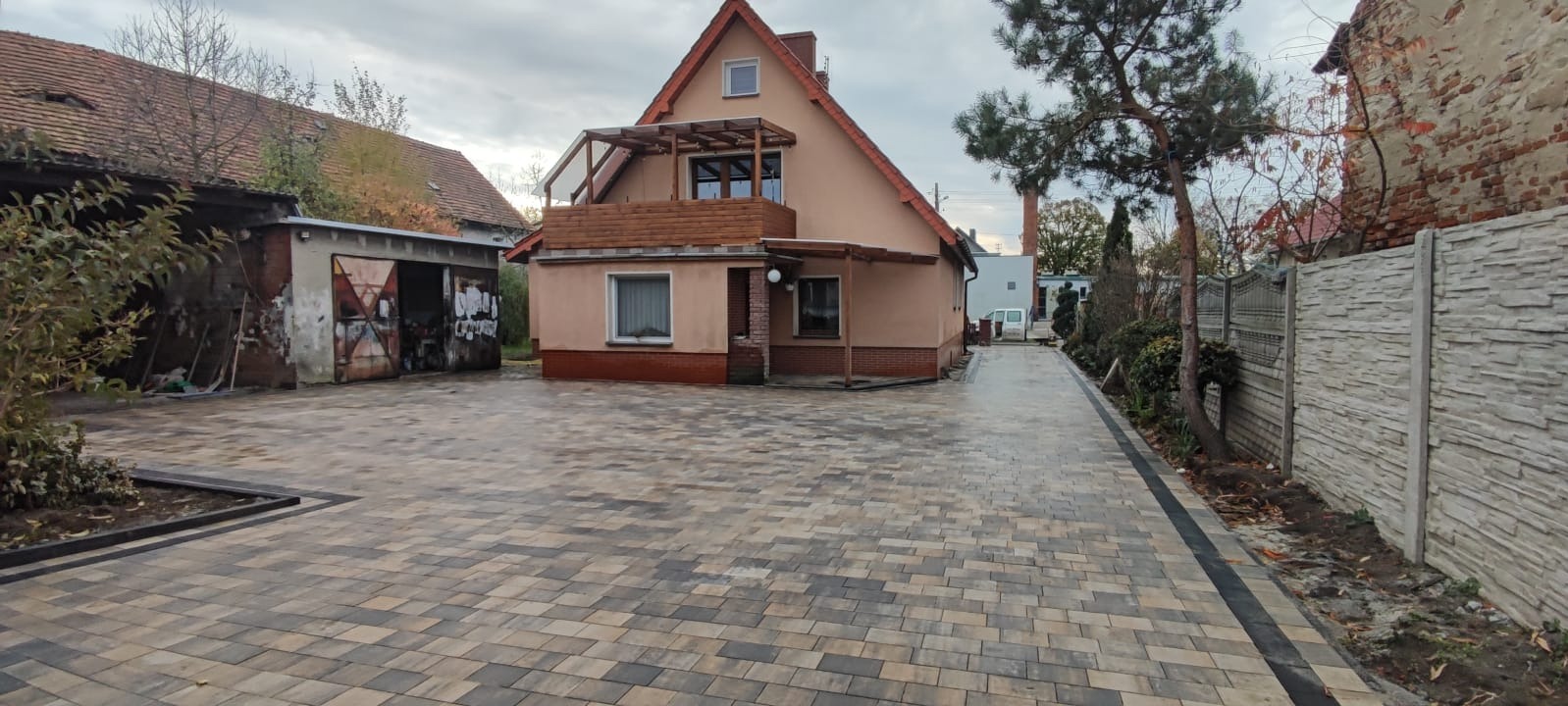 Ułożona kostka brukowa w odcieniach szarości i beżu prowadząca do domu z balkonem i garażem, widok na posesję przy pochmurnej pogodzie.