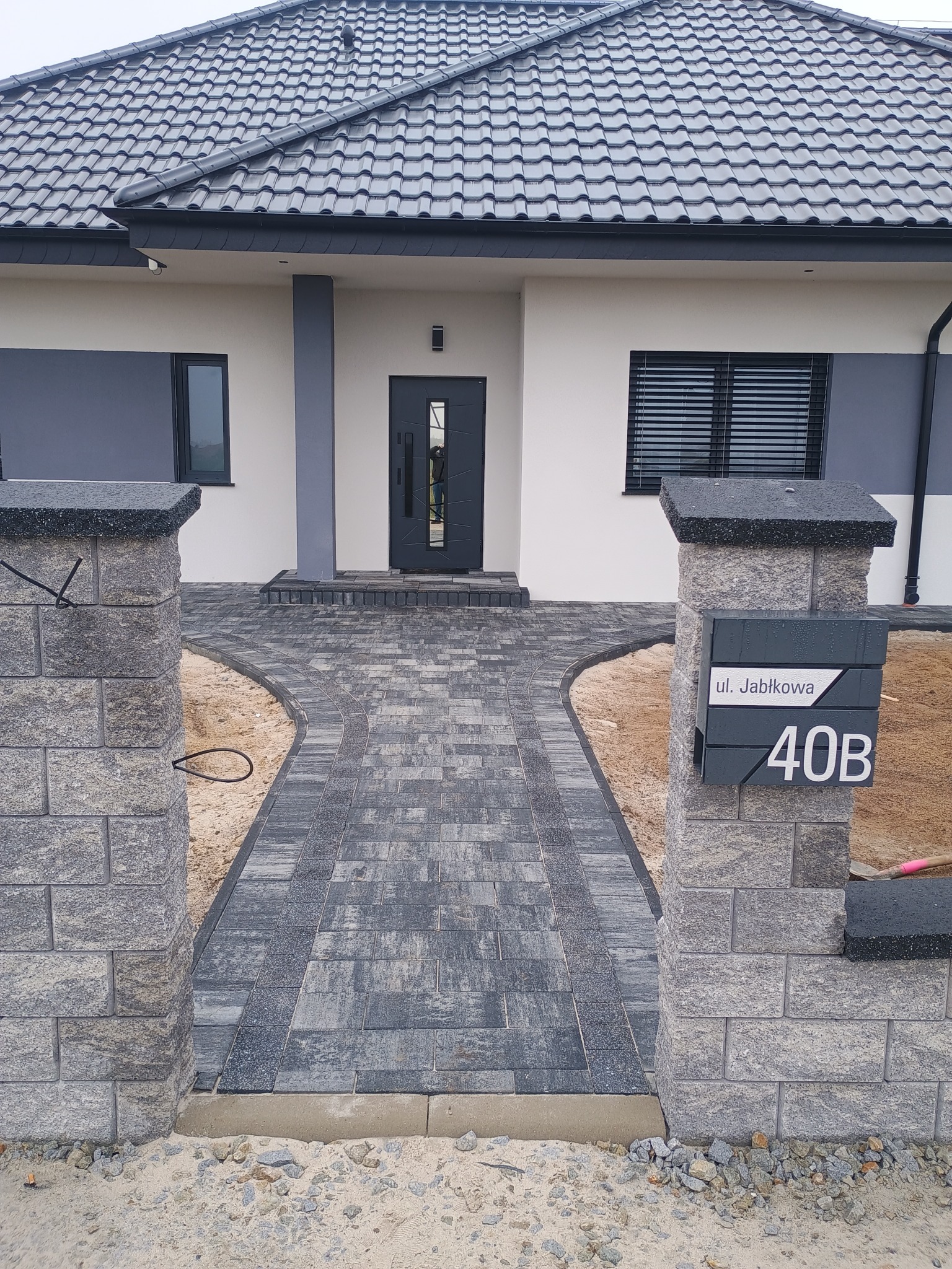 Nowoczesny dom z szarą kostką brukową prowadzącą do wejścia, widoczne słupki z kamienia z numerem domu '40B ul. Jabłkowa'.