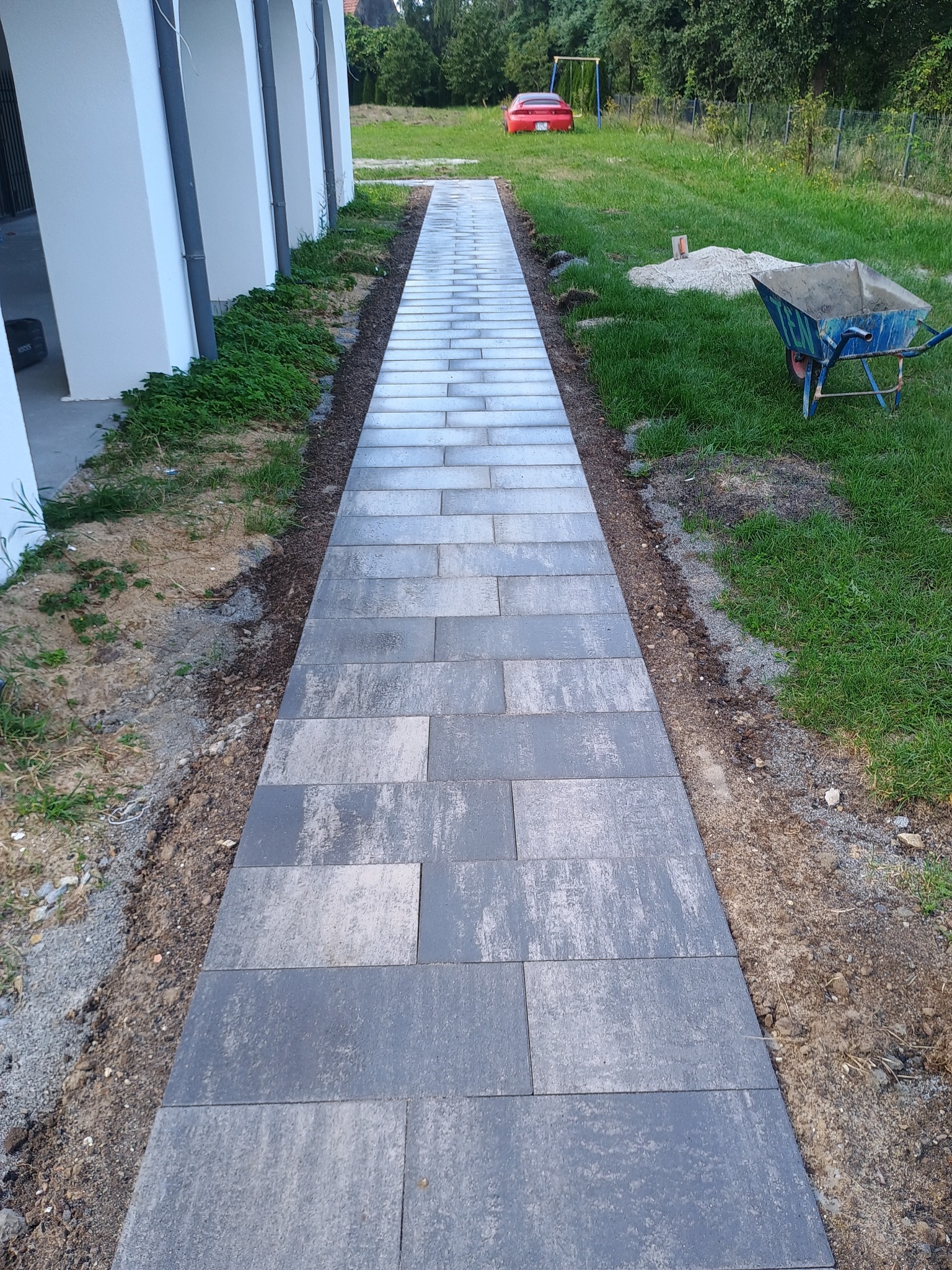 Ułożona ścieżka z szarych, prostokątnych płyt betonowych prowadząca przez trawnik, z widocznym w tle czerwonym samochodem i niebieską taczką.
