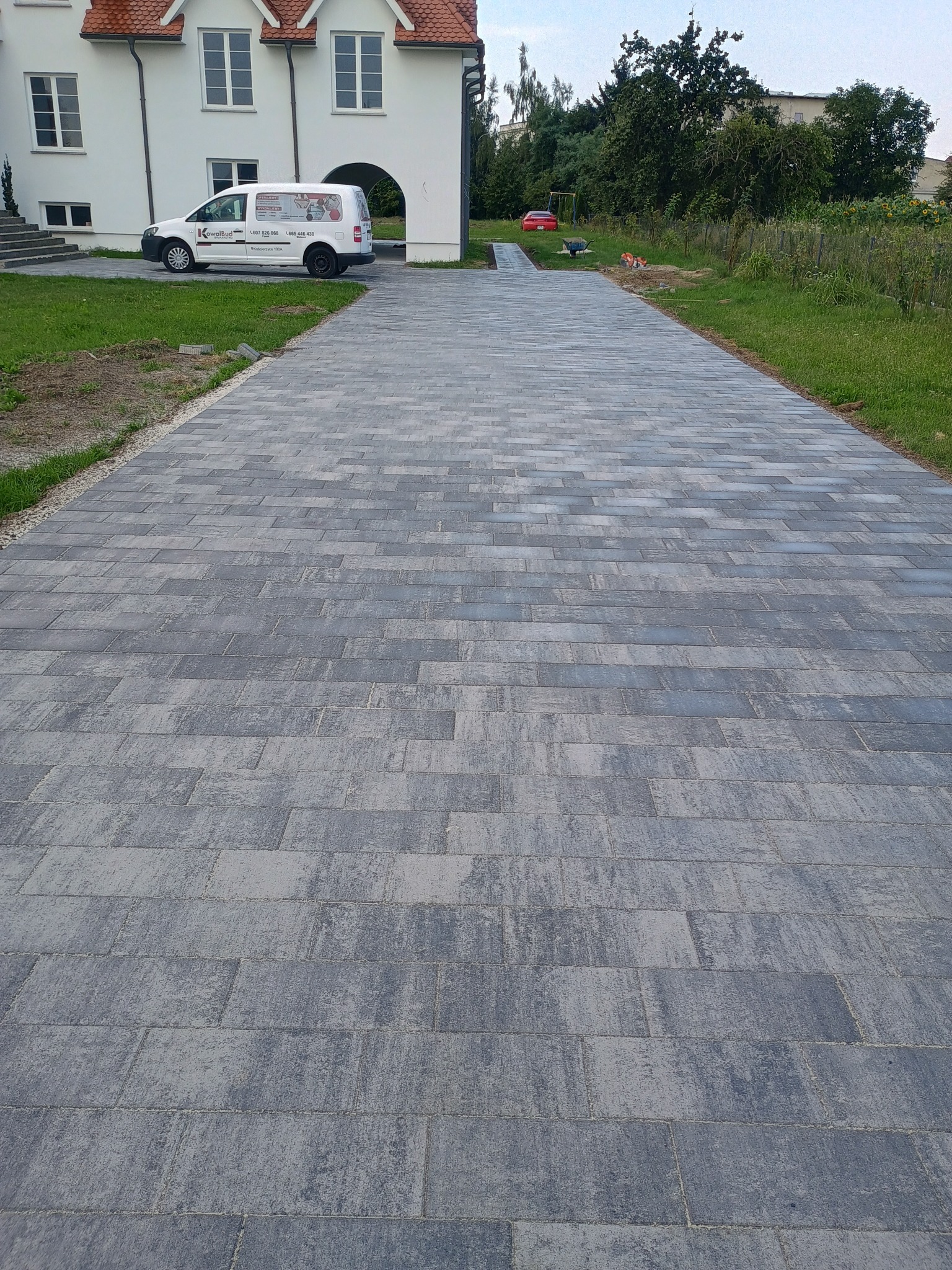 Długa, szara ścieżka z kostki brukowej prowadząca do białego domu z czerwonym dachem; w tle zaparkowany biały samochód dostawczy z logo firmy brukarskiej.