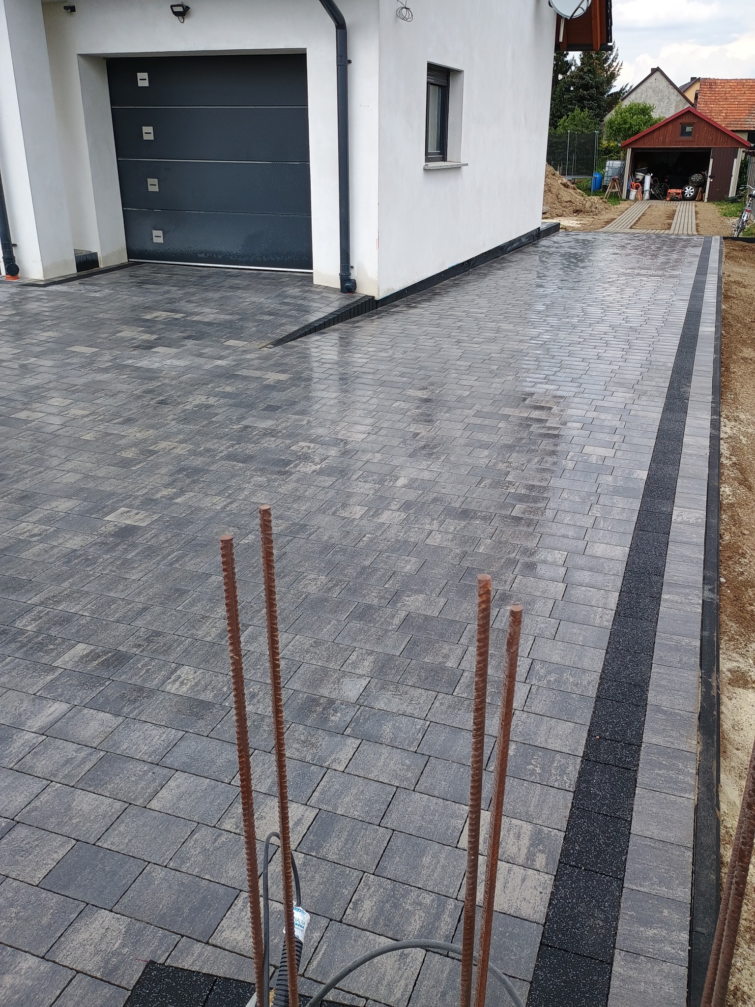 Świeżo ułożona, wilgotna kostka brukowa w odcieniach szarości przed garażem; widoczne pręty zbrojeniowe wskazujące na trwające prace wykończeniowe.