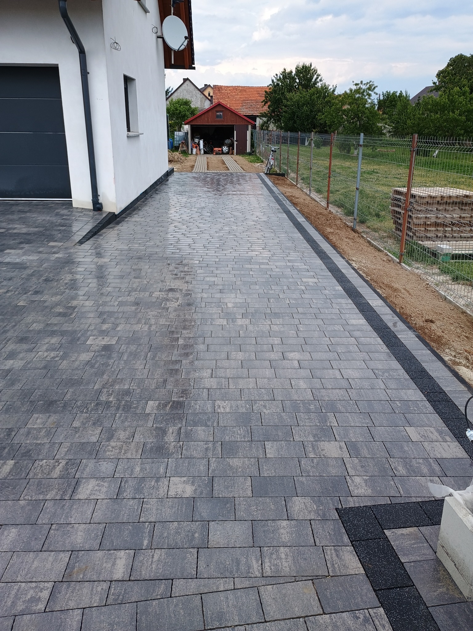 Długa, świeżo ułożona, szara kostka brukowa prowadząca od domu z garażem do oddalonego garażu, z widocznym odwodnieniem liniowym po bokach i fragmentem czarnej kostki przy krawędzi.