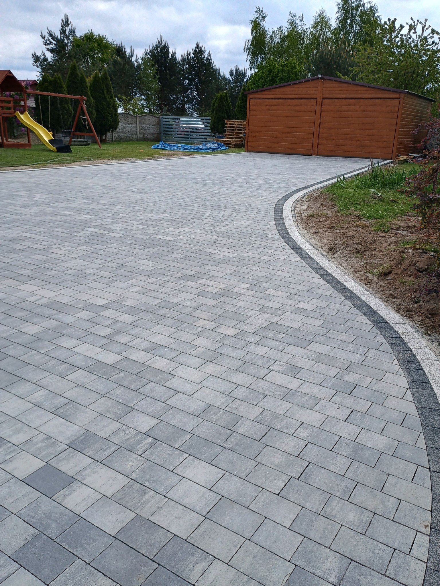 Ułożona szara kostka brukowa na podjeździe z obrzeżem z kostki granitowej, widoczny plac zabaw i garaż w tle.