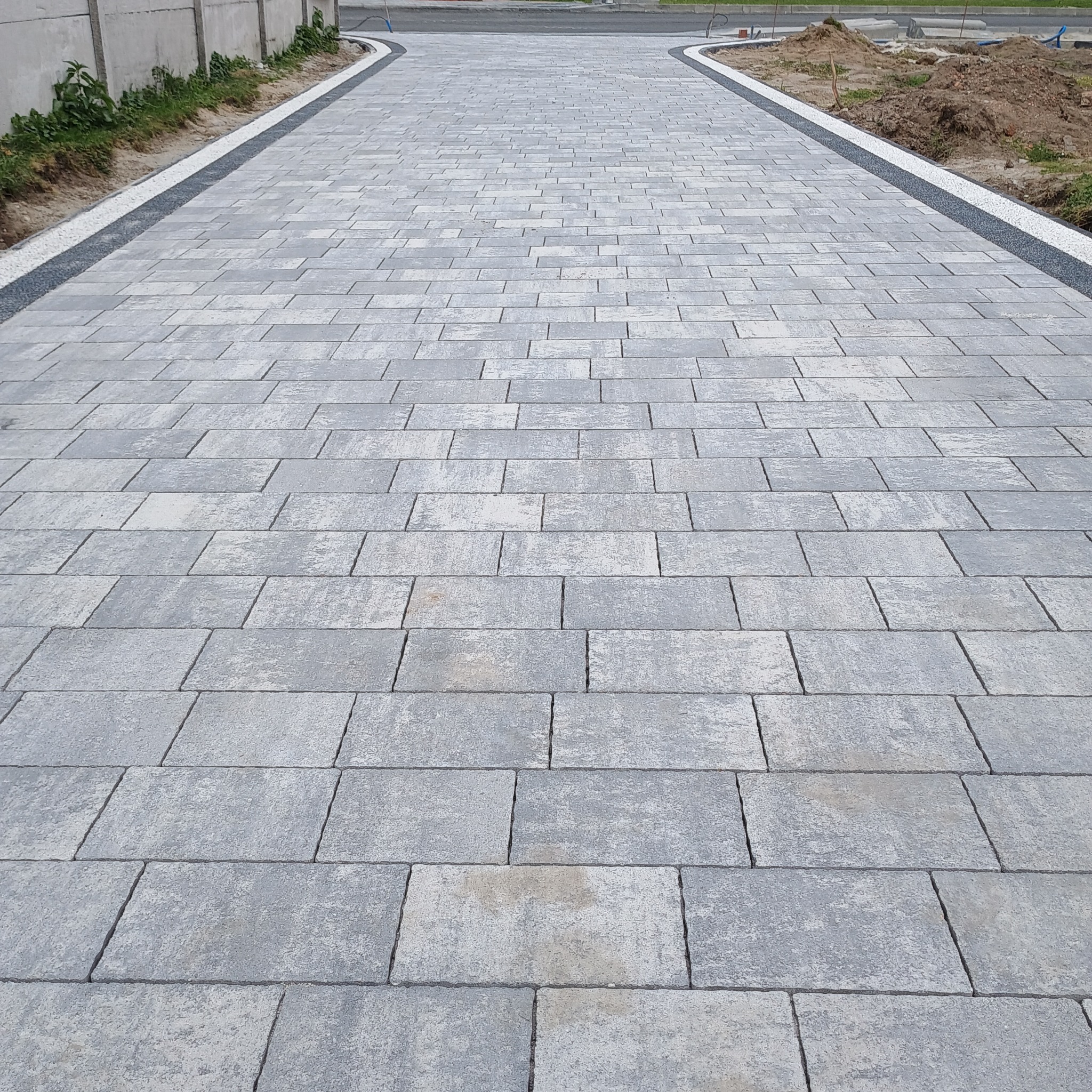 Nowo ułożona, długa, szara nawierzchnia z kostki brukowej w kształcie prostokątów, obrzeże z drobnego kamienia po obu stronach, widok w perspektywie.