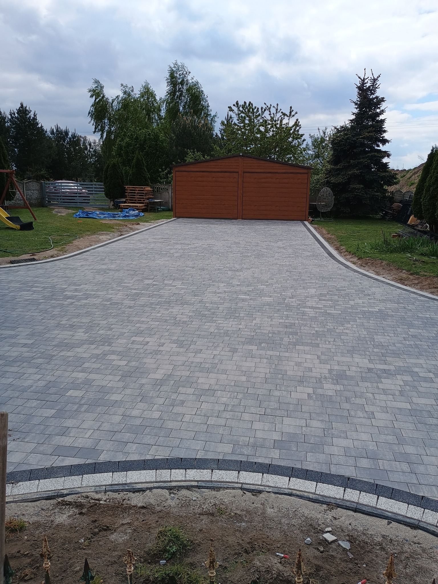 Szeroki podjazd z szarej kostki ułożonej we wzór jodełki, obrzeża z ciemnej kostki, w tle drewniany garaż i zieleń ogrodu, pochmurne niebo.