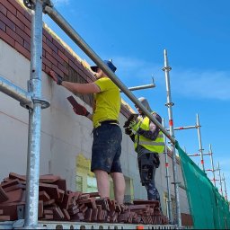 ANDRII HUBAL LED - Dwóch pracowników na rusztowaniu układa ceglane płytki elewacyjne na ścianie budynku, widoczne narzędzia i zapas płytek, błękitne niebo w tle.