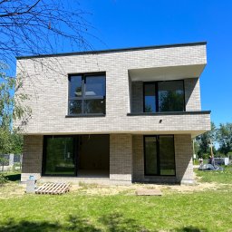 ANDRII HUBAL LED - Nowoczesny, dwukondygnacyjny dom z elewacją z jasnej cegły klinkierowej, czarne ramy okienne, teren wokół domu w trakcie prac wykończeniowych, paleta na trawniku.