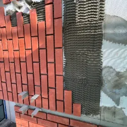 Montaż czerwonej cegły klinkierowej na elewacji budynku z widocznymi elementami mocującymi i nałożonym klejem w charakterystyczne fale.