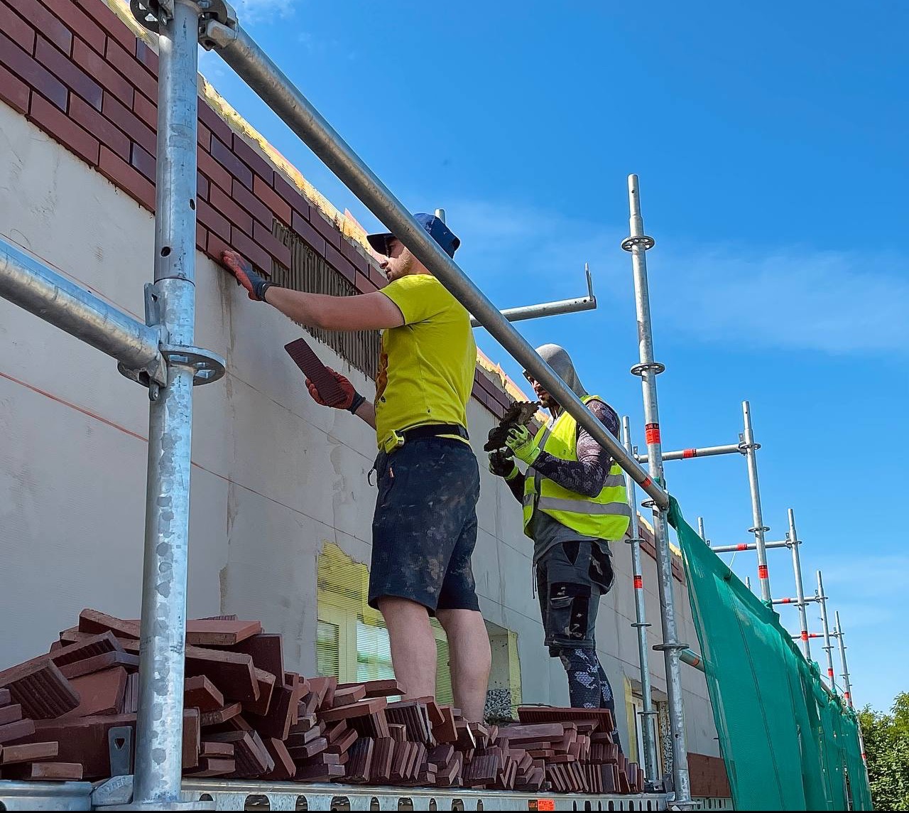 Dwóch pracowników na rusztowaniu układa ceglane płytki elewacyjne na ścianie budynku, widoczne narzędzia i zapas płytek, błękitne niebo w tle.
