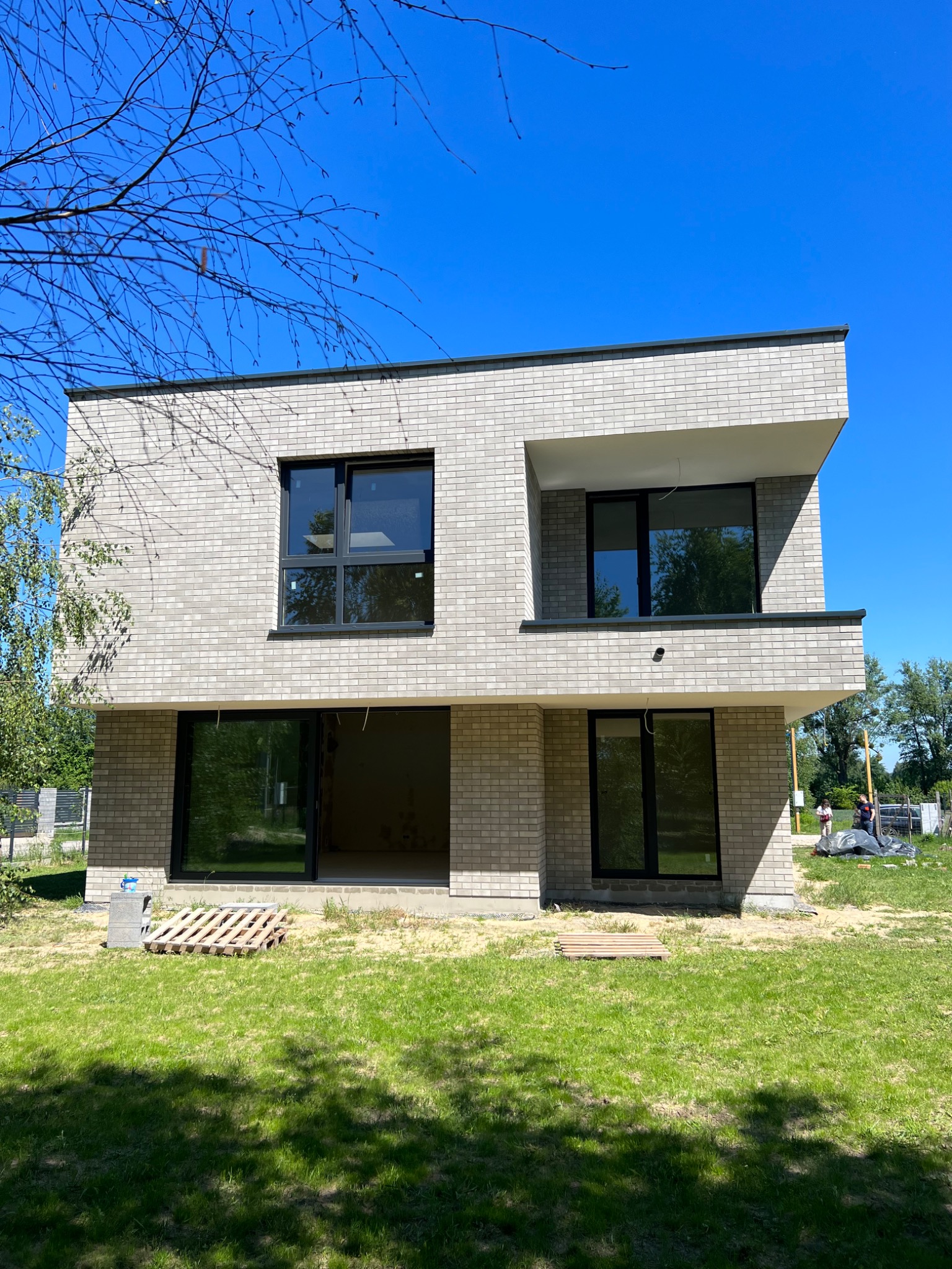 Nowoczesny, dwukondygnacyjny dom z elewacją z jasnej cegły klinkierowej, czarne ramy okienne, teren wokół domu w trakcie prac wykończeniowych, paleta na trawniku.