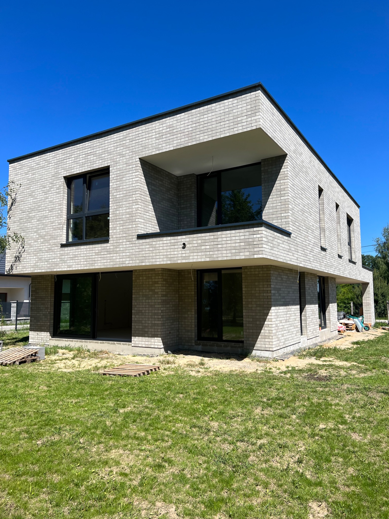 Nowoczesny dom z jasnoszarej cegły klinkierowej, widoczne czarne ramy okienne i balkon, trawnik w pierwszym planie, błękitne niebo w tle, teren wokół domu w trakcie prac wykończeniowych.
