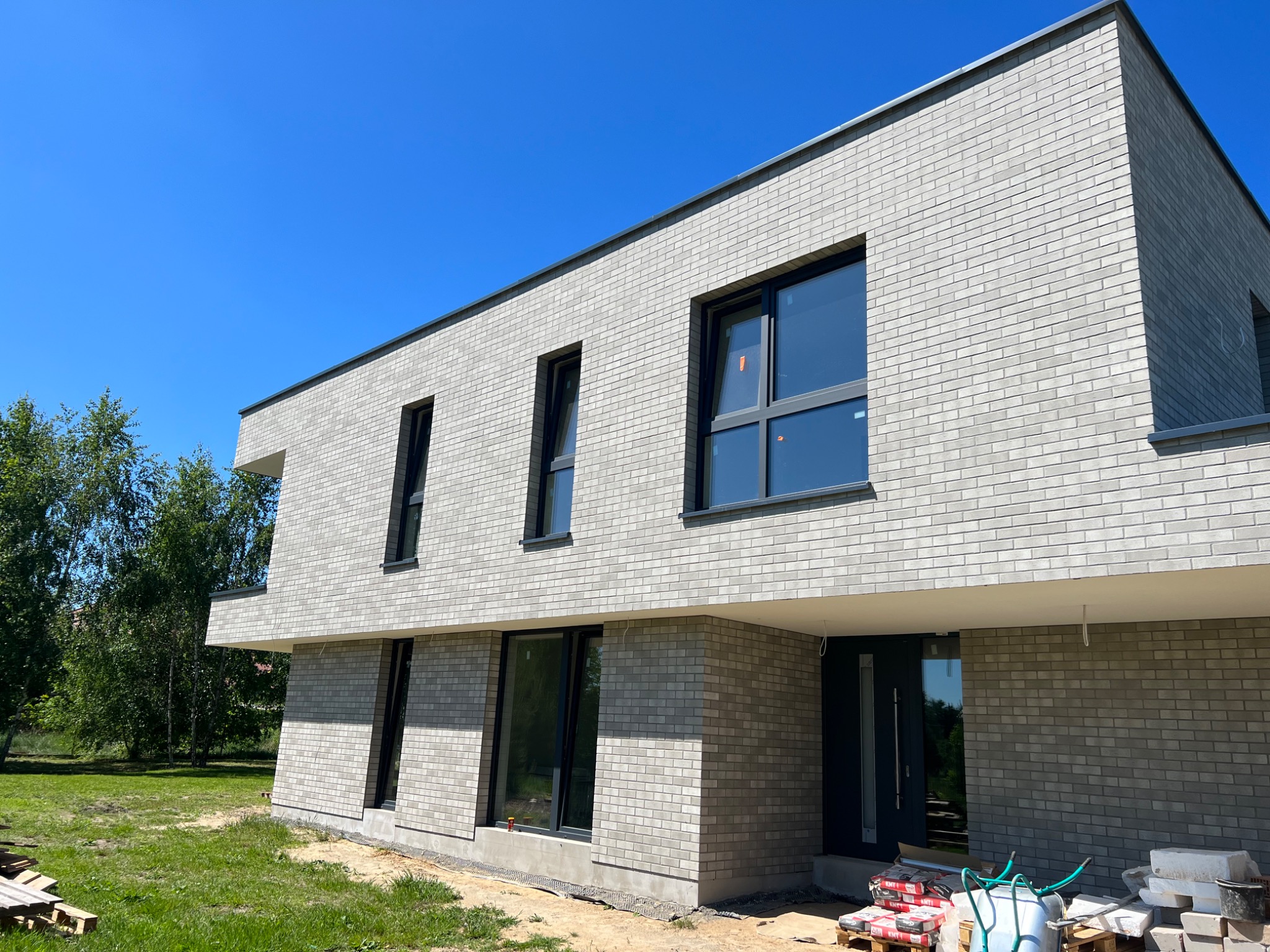 Nowoczesny dom z elewacją z jasnej cegły klinkierowej, czarne ramy okienne, teren wokół domu w trakcie zagospodarowywania, palety z materiałami budowlanymi przy wejściu.