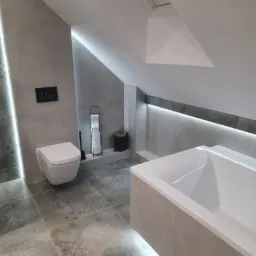 Nowoczesna łazienka na poddaszu z podświetleniem LED, wiszącą toaletą i wanną obudowaną płytkami w odcieniach szarości oraz wzorzystą podłogą.