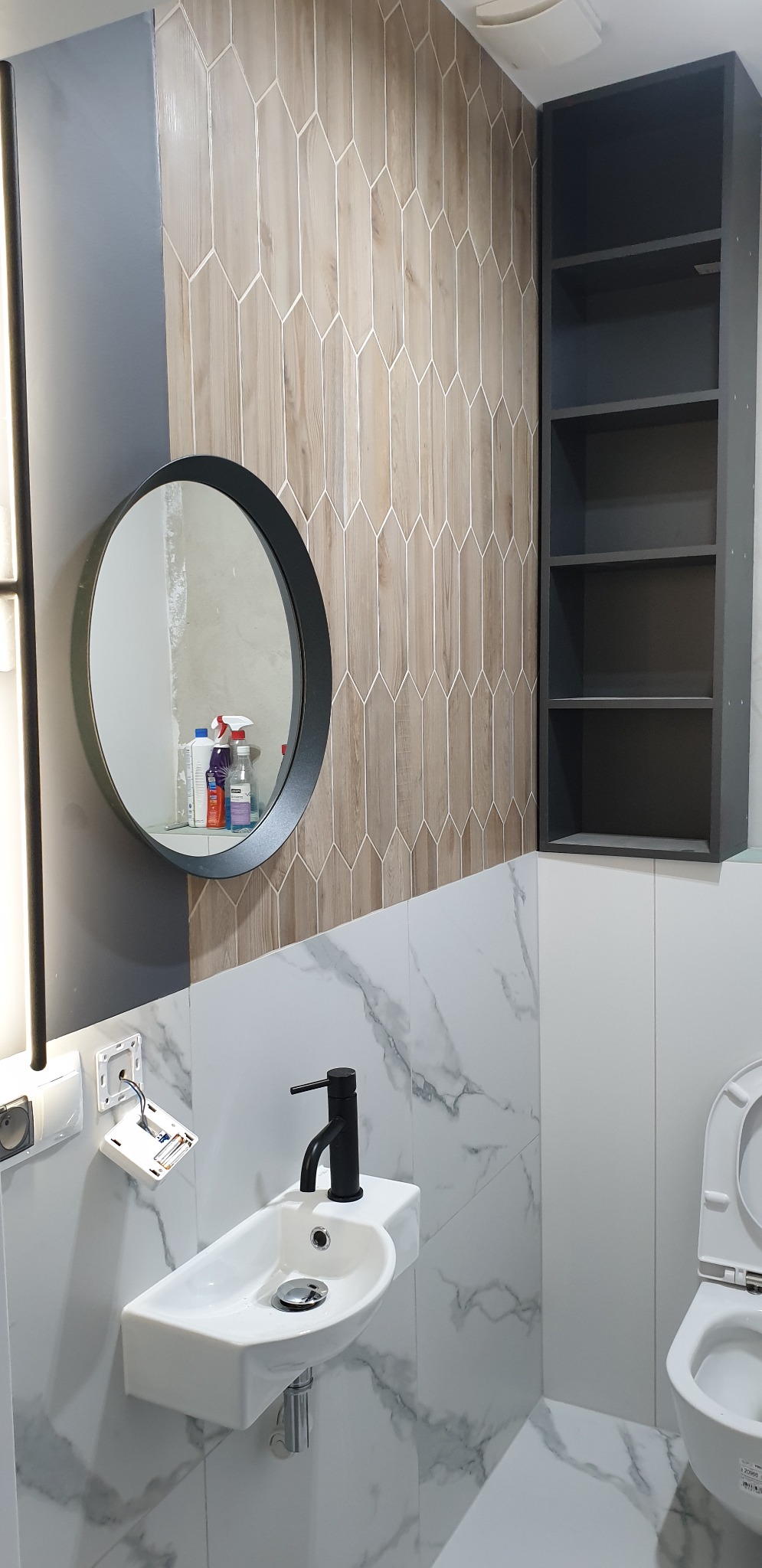 Umywalka nablatowa z czarną armaturą na tle ściany z białych płytek imitujących marmur i drewnopodobnych płytek w kształcie rombów, obok toaleta i czarny regał wiszący.