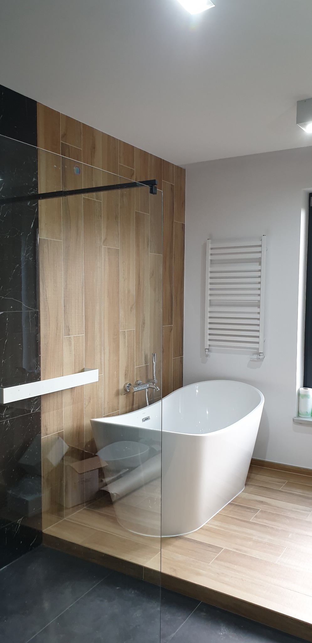 Nowoczesna łazienka z wolnostojącą białą wanną, szklaną kabiną prysznicową z czarnymi elementami, grzejnikiem drabinkowym i drewnopodobnymi płytkami na ścianach.