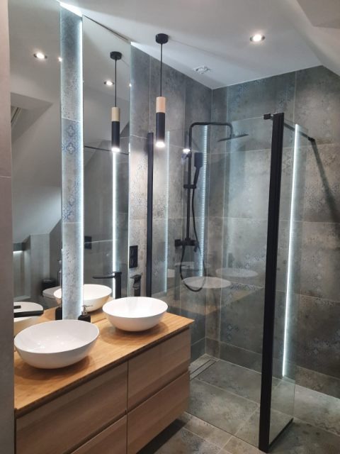 Nowoczesna łazienka z dwoma okrągłymi umywalkami nablatowymi na drewnianej szafce, czarną armaturą i kabiną prysznicową typu walk-in z czarnymi profilami, oświetlona lampami wiszącymi z drewnianymi...