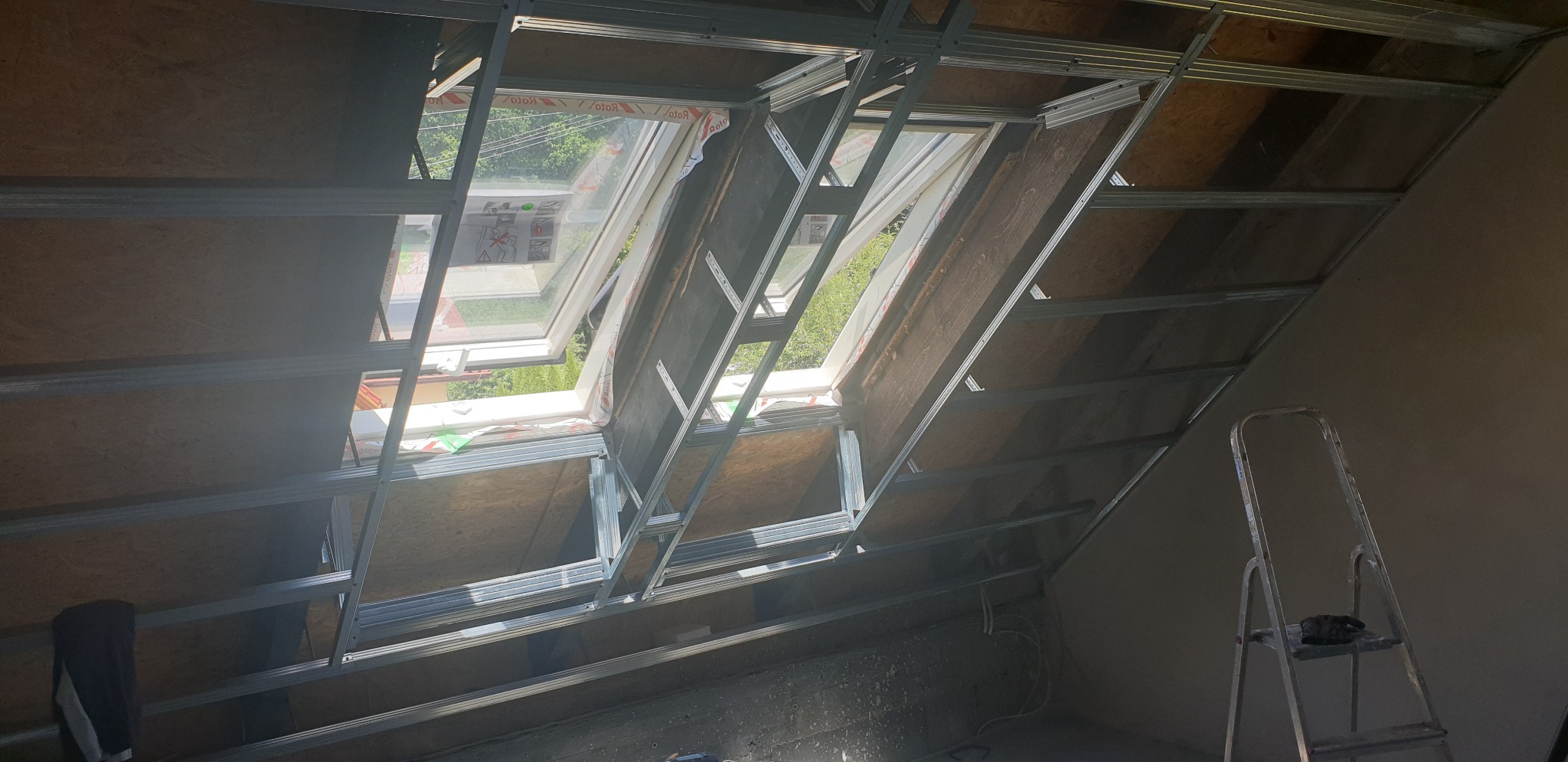 Montaż metalowej konstrukcji pod sufitem skośnym na poddaszu, widoczne okna dachowe i drabina oparta o ścianę.