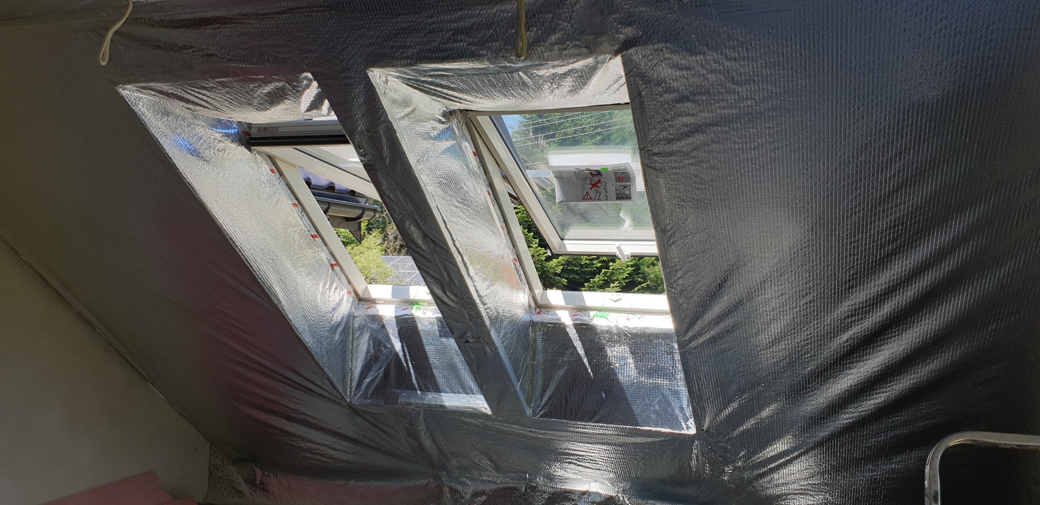 Montaż okien dachowych z folią termoizolacyjną, widok z wnętrza poddasza na dwa otwarte okna z widokiem na zieleń.